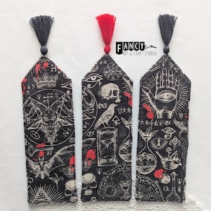 Puede incluir: Tres marcapáginas de tela negra con diseños rojos y blancos. Los marcapáginas presentan varios símbolos ocultos, incluyendo una calavera, un ojo, una mano, un murciélago y un pentagrama. Los marcapáginas tienen borlas negras.