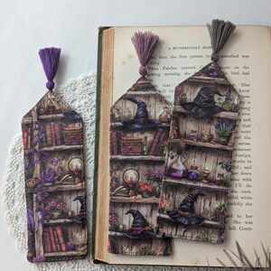 May include: Three decorative bookmarks with a witchy theme. Each bookmark features a detailed illustration of a bookshelf with books, potions, and a witch's hat. The bookmarks have purple tassels and are placed on a book.