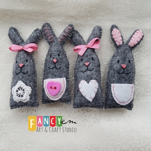 May include: Four gray felt bunny finger puppets with pink accents. Each bunny has a different detail: a white flower, a pink button, a white heart, and a white pocket. The bunnies are arranged in a row on a white fabric background. The text "FANCYem ART & CRAFT STUDIO" is visible below the bunnies.