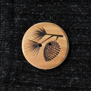 May include: A round, light-colored wooden button with a detailed engraved design of a pine branch with needles and a pinecone. The button has two holes and is set against a dark gray, textured fabric background.