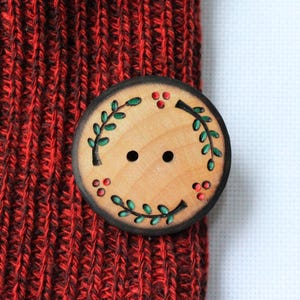 May include: A round wooden button with two holes, featuring a decorative design of green leaves and red berries around the edge. The button is attached to a red knitted fabric.