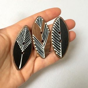 May include: Three wooden buttons with a black and white geometric pattern. The buttons are oval shaped and have two holes each.