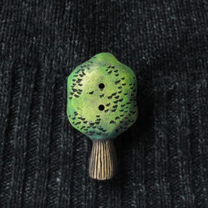 May include: A green tree-shaped button with two holes, set against a dark gray textured background. The button has a textured surface with black detailing and a brown, vertical-lined base. This unique button adds a whimsical touch to any sewing project.