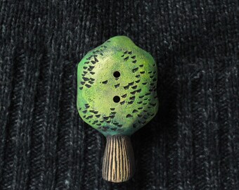 May include: A green tree-shaped button with two holes, set against a dark grey textured background. The button has a textured surface with black detailing and a brown, vertically-lined base. This unique button adds a whimsical touch to any sewing project.