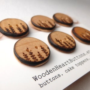 May include: Six wooden buttons with a burnt design of a plant. The buttons are arranged in a row on a white card with the text "WoodenHeartButtons. buttons. cake toppers."