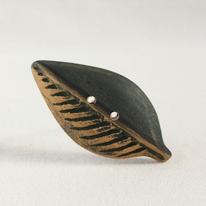 May include: A leaf-shaped button featuring a dark charcoal gray surface with textured, recessed bronze-colored veins. The button includes two small, circular holes positioned centrally for attachment, showcasing a distinct contrast between the dark matte finish and the metallic bronze detailing.