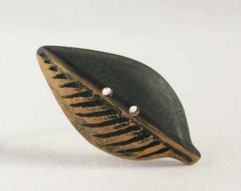 May include: A leaf-shaped button featuring a dark charcoal grey surface with textured, recessed bronze-coloured veins. The button includes two small, circular holes positioned centrally for attachment, showcasing a distinct contrast between the dark matte finish and the metallic bronze detailing.
