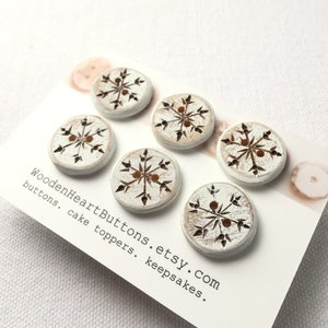 May include: Six white wooden buttons with a brown snowflake design. The buttons are arranged in a circle on a white card with the text "WoodenHeartButtons.etsy.com buttons. cake toppers. keepsakes."
