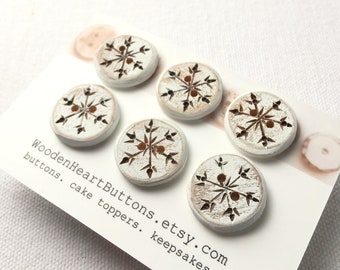 May include: Six white wooden buttons with a brown snowflake design. The buttons are arranged in a circle on a white card with the text "WoodenHeartButtons.etsy.com buttons. cake toppers. keepsakes."