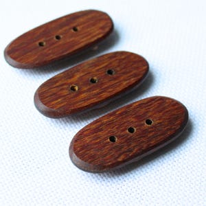 May include: Three oval-shaped wooden buttons with three holes each. The buttons are a rich, dark brown color with visible wood grain. The buttons are arranged diagonally against a white background.