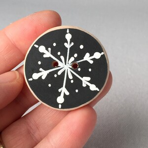 May include: A black wooden button with a white snowflake design. The snowflake has six points and is surrounded by small white dots.