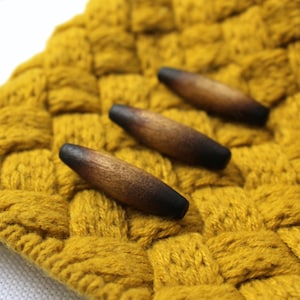 May include: Three elongated, spindle-shaped wooden toggles with a smooth, polished finish. Each toggle features a natural wood grain that transitions from a light brown center to dark, charred-looking tips at both ends, arranged diagonally.