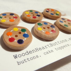 May include: Six wooden buttons with a rainbow circle design. The buttons are painted with red, orange, yellow, green, blue, and purple dots. The text "WoodenHeartButtons. buttons. cake toppers." is printed on the white surface below the buttons.