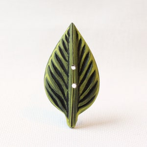 May include: A leaf-shaped button with a green and black painted design. The button has two small holes for sewing and is made of wood. The button is a decorative element for clothing or crafts.