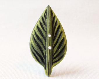 May include: A leaf-shaped button with a green and black painted design. The button has two small holes for sewing and is made of wood. The button is a decorative element for clothing or crafts.
