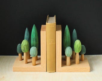 May include: Wooden bookends with a forest scene. The bookends are made of light-colored wood and feature a row of carved trees in shades of green. A book with the title "Man & Superman" stands between the bookends.