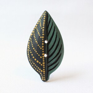 May include: A leaf-shaped wooden button. One half is dark green with carved veins, the other is black with a dotted gold pattern. Two small holes are visible for sewing. The button is designed to look like a leaf.