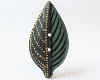 May include: A leaf-shaped wooden button. One half is dark green with carved veins, the other is black with a dotted gold pattern. Two small holes are visible for sewing. The button is designed to look like a leaf.