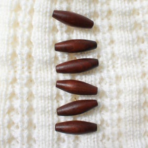 May include: A set of six dark brown, elongated, spindle-shaped wooden toggle buttons arranged in a vertical column. Each button features a smooth, polished finish with visible wood grain patterns and a small metal shank loop on the underside.