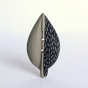 May include: A leaf-shaped ceramic button with a split design. One half is a muted gray, the other is dark blue with white dashes. Two small, copper-colored rivets are visible.