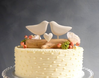 May include: A white wedding cake with a white buttercream frosting and a dotted pattern. The cake is decorated with a wooden log, two white birds, and a small white heart.