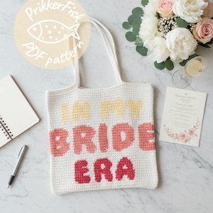 May include: White crocheted tote bag with the text "IN MY BRIDE ERA" in yellow, coral, and red. The bag is next to a bouquet of flowers, a notebook, a pen, and a wedding invitation.
