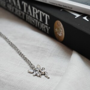 Peut inclure: Un collier en cha&icirc;ne argent&eacute;e avec un pendentif en forme de f&eacute;e. La f&eacute;e est repr&eacute;sent&eacute;e avec des ailes d&eacute;ploy&eacute;es et une robe flottante. Le collier est pos&eacute; sur une surface en tissu blanc. Le livre "The Secret History" de Donna Tartt est visible en arri&egrave;re-plan.