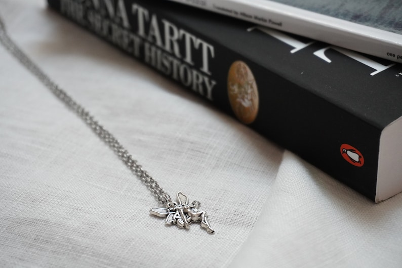 Peut inclure: Un collier en cha&icirc;ne argent&eacute;e avec un pendentif en forme de f&eacute;e. La f&eacute;e est repr&eacute;sent&eacute;e avec des ailes d&eacute;ploy&eacute;es et une robe flottante. Le collier est pos&eacute; sur une surface en tissu blanc. Le livre "The Secret History" de Donna Tartt est visible en arri&egrave;re-plan.