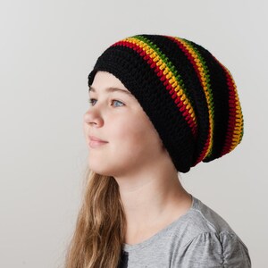 Op de afbeelding: Een jong persoon draagt ​​een zwarte beanie met rode, gele en groene strepen. De beanie is gebreid en heeft een slouchy pasvorm.