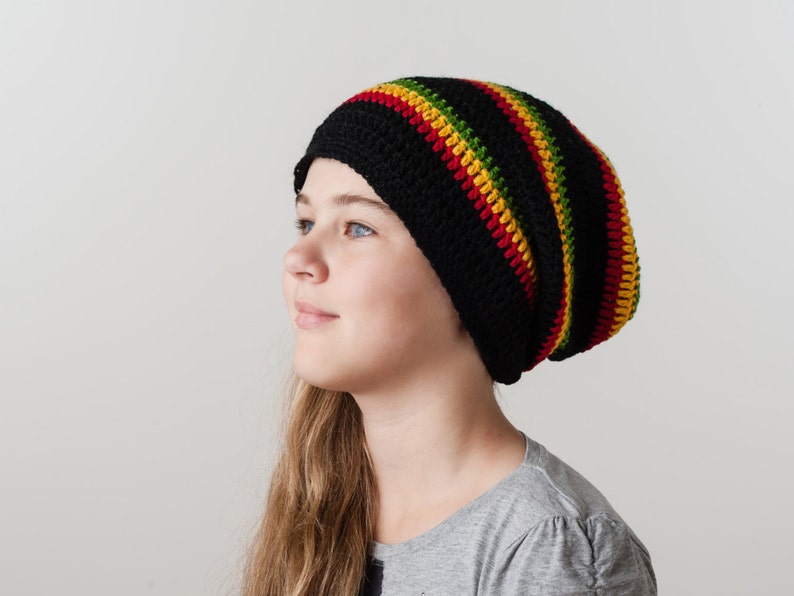 Op de afbeelding: Een jong persoon draagt ​​een zwarte beanie met rode, gele en groene strepen. De beanie is gebreid en heeft een slouchy pasvorm.