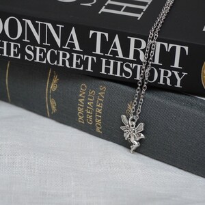 Peut inclure: Un collier avec un pendentif en forme de f&eacute;e argent&eacute; pos&eacute; sur deux livres reli&eacute;s en noir. Le livre du dessus est "Donna Tartt The Secret History" et le livre du dessous est "Dorian Gray" d'Oscar Wilde.