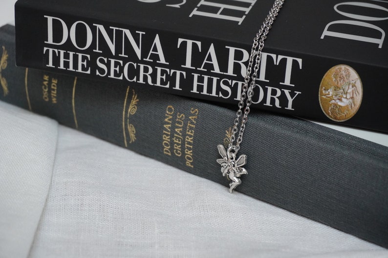 Peut inclure: Un collier avec un pendentif en forme de f&eacute;e argent&eacute; pos&eacute; sur deux livres reli&eacute;s en noir. Le livre du dessus est "Donna Tartt The Secret History" et le livre du dessous est "Dorian Gray" d'Oscar Wilde.