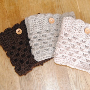 Op de afbeelding: Drie gebreide beenwarmers met een knoopdetail. De beenwarmers zijn in bruin, beige en lichtbeige.