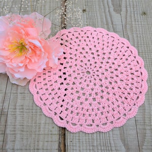 May include: A pink crocheted doily with a delicate, intricate design. The doily is circular and has a scalloped edge. It is lying on a wooden surface.