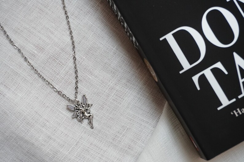 Peut inclure: Un collier en cha&icirc;ne argent avec un pendentif en forme de f&eacute;e. La f&eacute;e est repr&eacute;sent&eacute;e avec des ailes et une robe flottante.