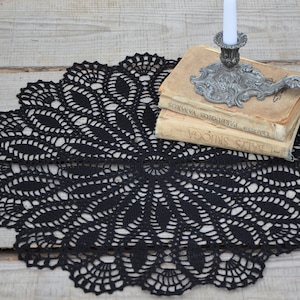 May include: Black crochet doily with a delicate, intricate design. A silver candlestick with a white candle sits on top of a stack of old books.
