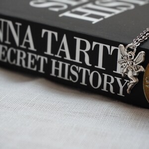 Peut inclure: Un livre noir avec le titre "DONNA TARTT THE SECRET HISTORY" sur la couverture. Un charme de f&eacute;e argent&eacute; pend &agrave; une cha&icirc;ne argent&eacute;e sur le livre.