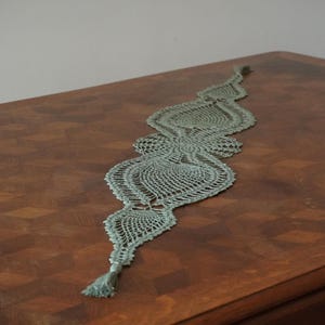 May include: A light green crocheted table runner with a scalloped design and a tassel at the end. The runner is on a wooden table.