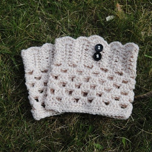 May include: A pair of crocheted leg warmers in a light beige color with a black button on the top. The leg warmers are on a bed of green grass.