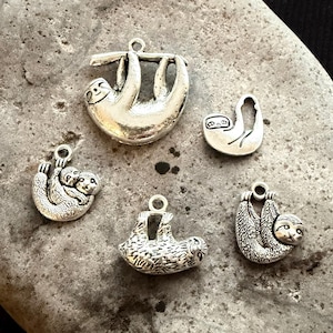 May include: A collection of five silver-toned metal sloth charms in various poses. Each charm features a small loop at the top for attachment. The designs include a large hanging sloth, a small outline sloth, a mother and baby sloth, and two others.