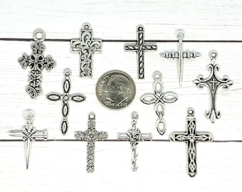 May include: A collection of 12 silver-toned cross charms with various designs, including floral, ornate, and simple. The charms are arranged on a white wooden surface.
