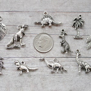 May include: A collection of silver charm pendants featuring various dinosaur designs, palm trees, and leaves. The charms are approximately the size of a US quarter.