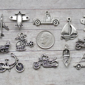 May include: A collection of 13 silver charm pendants featuring various transportation themes, including a plane, helicopter, truck, sailboat, motorcycle, bicycle, and car. The charms are displayed on a wooden surface.