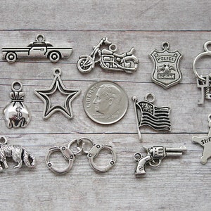 May include: A collection of silver-toned metal charms featuring various law enforcement and crime-related themes. The charms include a police car, motorcycle, police badge, handcuffs, a gun, a bag of money, a bullet, a star, a sheriff's star, and a key.