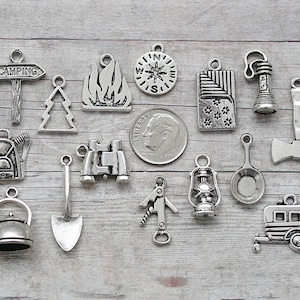 May include: A collection of silver-toned metal charms with camping and outdoor themes. The charms include a compass, a campfire, a tree, a lantern, a backpack, binoculars, a shovel, a kettle, a frying pan, an axe, a flashlight, a sign that says "Camping", and a camper trailer.