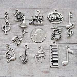 May include: A collection of silver-toned metal charms featuring musical instruments and notes. The charms include a violin, treble clef, french horn, saxophone, guitar, piano, drum set, flute, trumpet, and music notes. The charms are arranged on a wooden surface. "I Love Music" is written on one of the charms.