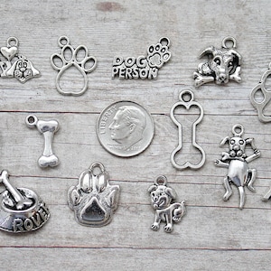 May include: A collection of 15 silver-toned metal charms with dog-themed designs. The charms include paw prints, bones, dog silhouettes, a dog house, a fire hydrant, and a dog bowl. The charms are perfect for adding a touch of personality to jewelry, keychains, or other crafts.