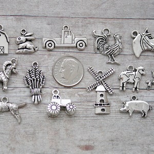 May include: A collection of 14 silver-toned metal charms featuring farm animals, a barn, a tractor, a windmill, a scarecrow, and wheat. The charms are arranged on a wooden surface.