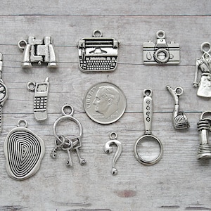 May include: A collection of silver-toned charms with detective-themed designs. Includes a watch, binoculars, typewriter, camera, trench coat, fingerprint, keys, question mark, magnifying glass, and a phone.