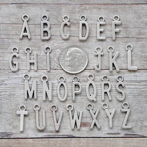 May include: A set of 26 silver metal alphabet letter charms with a jump ring. The charms are arranged in alphabetical order on a wooden surface. The charms are approximately the size of a US dime.